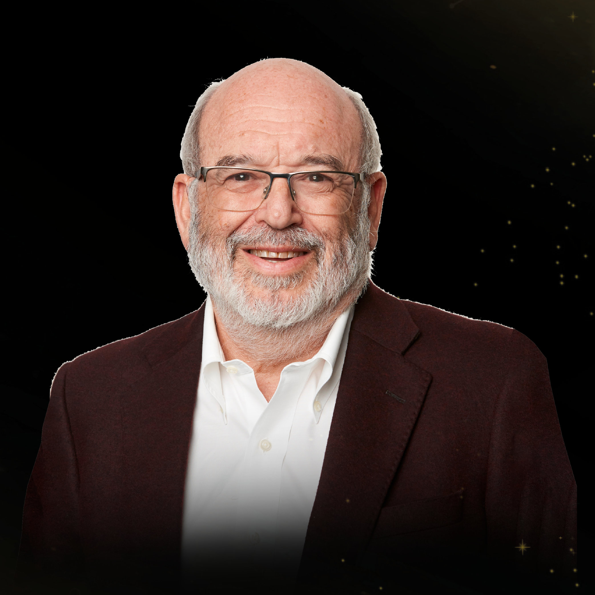 Older bald man with a gray beard and glasses, smiling, wearing a dark red blazer and white shirt on a black background.
